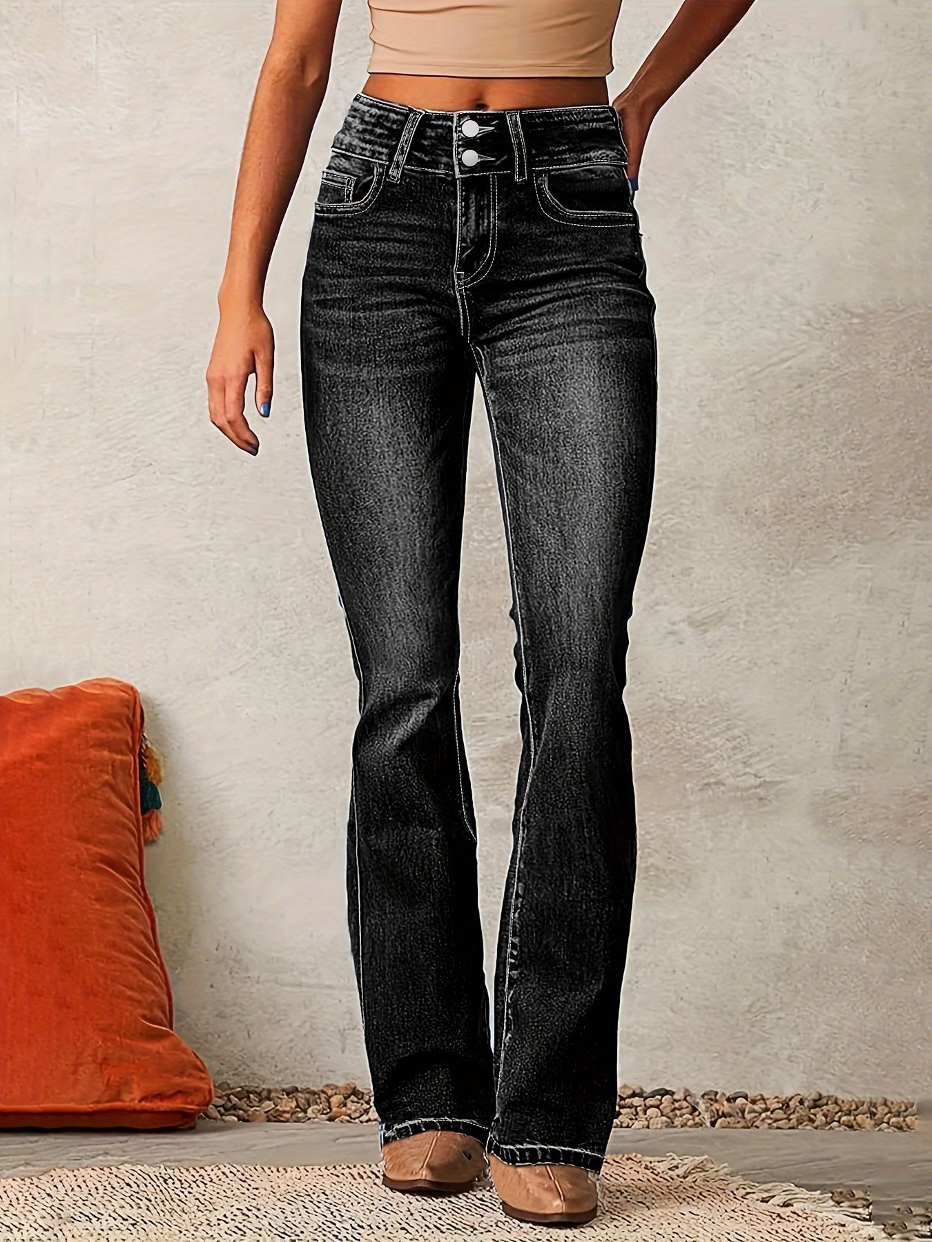 Person wearing black jeans standing against a neutral wall with an orange pillow on the floor.