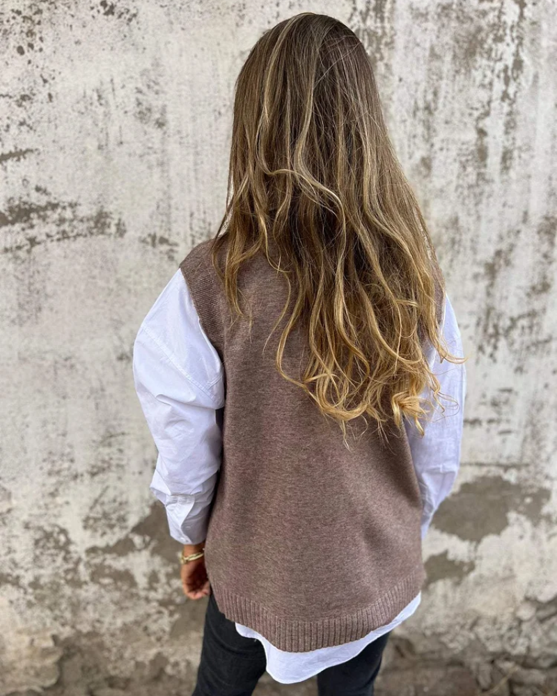 Person wearing a brown knitted vest over a white shirt against a textured wall.