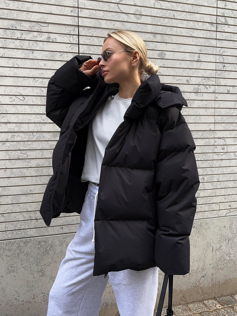 Person wearing a black puffer jacket and white pants against a gray wall.