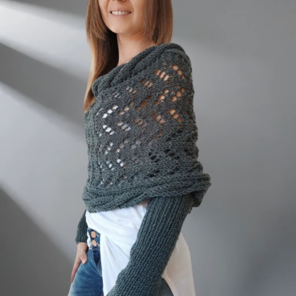 Woman wearing a dark green knitted shawl over a white shirt and jeans on a plain background