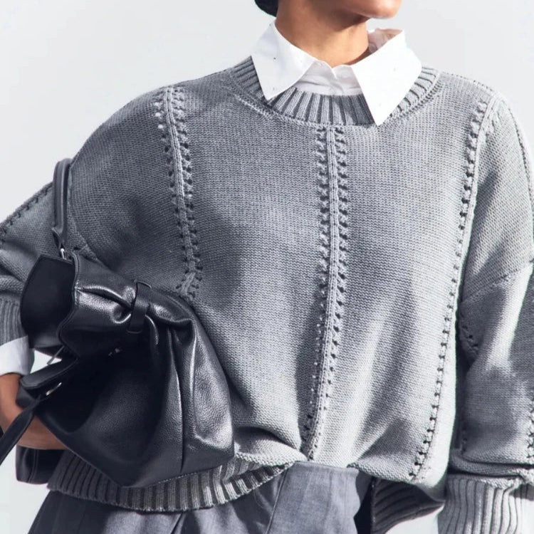 Person wearing a gray sweater with a white shirt underneath, holding a black bag against a light background