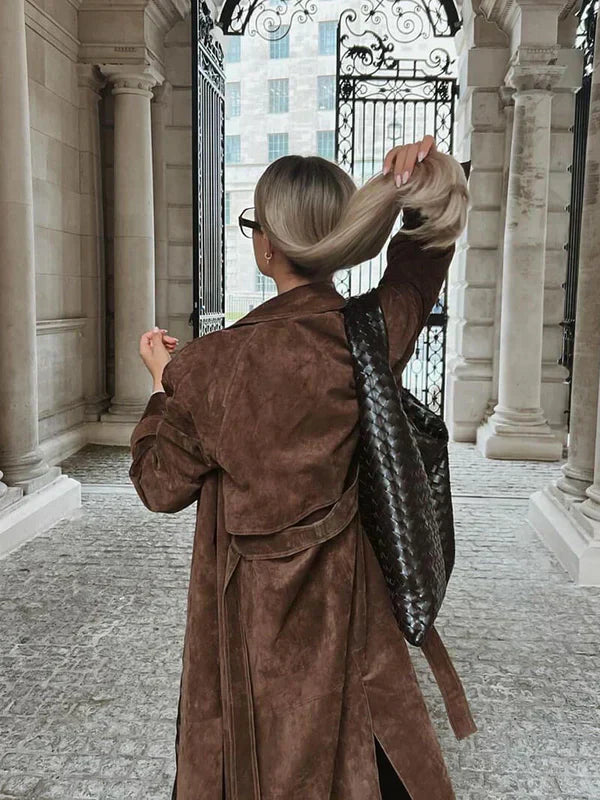 Person in a brown coat with a patterned backpack standing in an elegant outdoor setting.