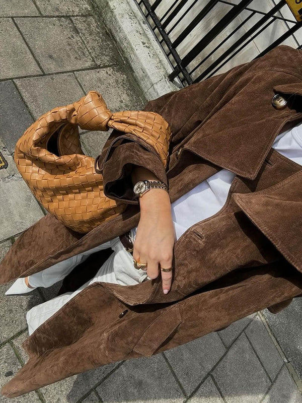 Person wearing a brown suede coat with a woven handbag on a sidewalk.