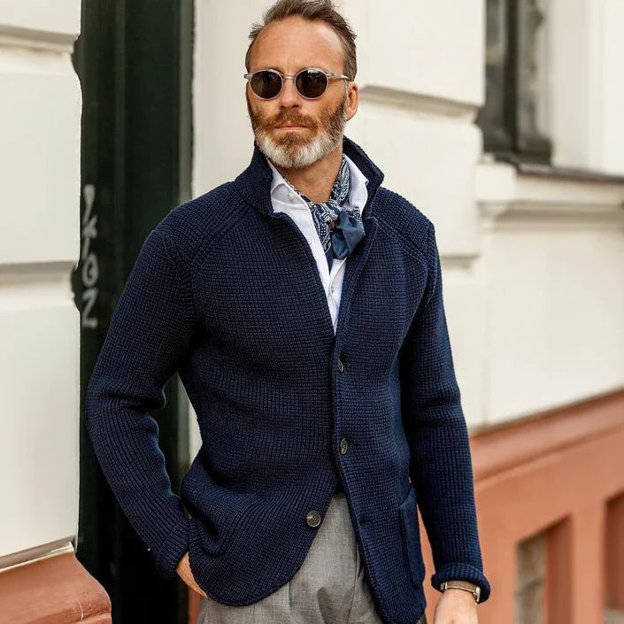 Man wearing a navy blue cardigan over a light shirt with sunglasses, standing against a building.