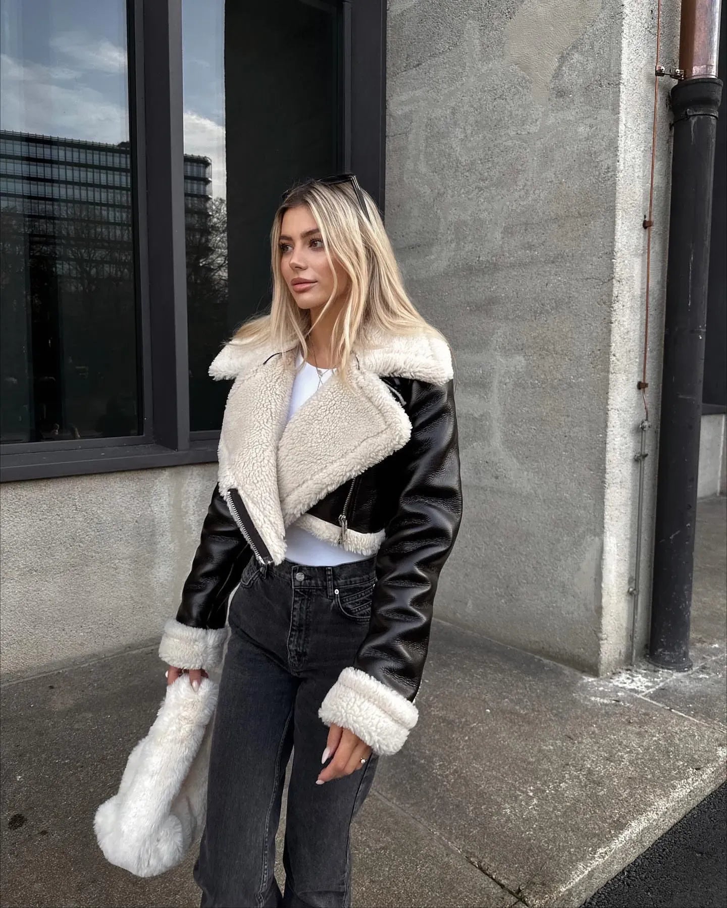 Woman wearing a black leather jacket with white fur trim standing on a sidewalk.