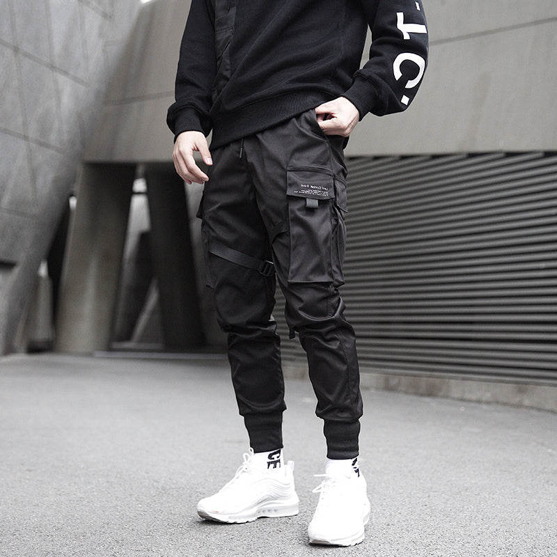 Person wearing black cargo pants and a black jacket with white text, standing in an urban setting.