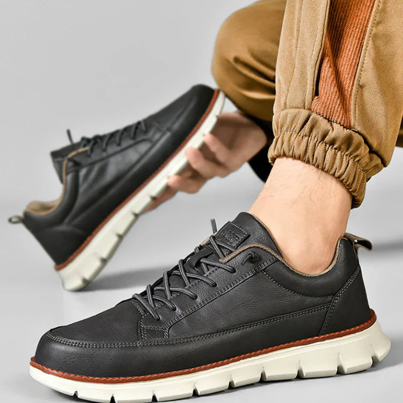 Black leather shoes with white soles worn by a person in brown pants on a gray background