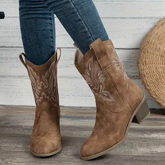 Brown cowboy boots with intricate designs worn by a person on a wooden floor.