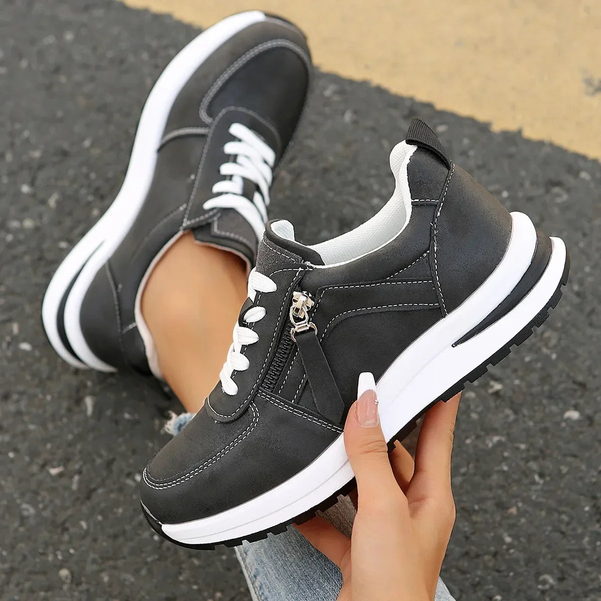 Black sneakers with white laces and sole held by a hand on a pavement background