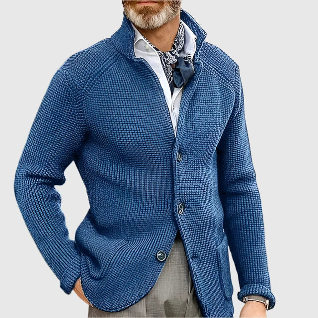 Man wearing a blue knitted cardigan over a white shirt with a patterned tie on a light gray background