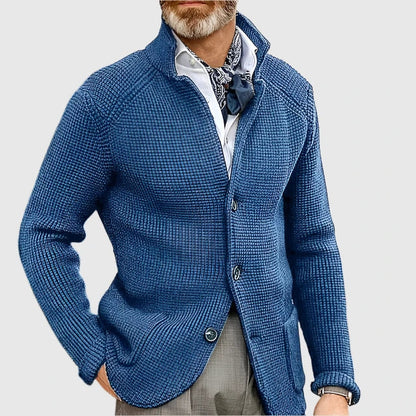 Man wearing a blue knitted cardigan over a white shirt with a patterned tie on a light gray background