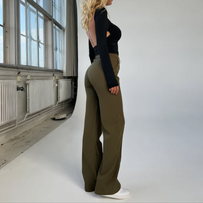Person wearing a black top and green pants standing in a room with large windows.